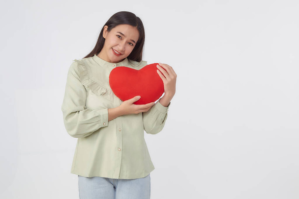 Piękno młoda brunetka Azji kobieta noszenie zielonej koszuli stojące trzymać czerwony serce kształt poduszki w jasnoszary odizolowane studio tło, studio portret - Zdjęcie, obraz