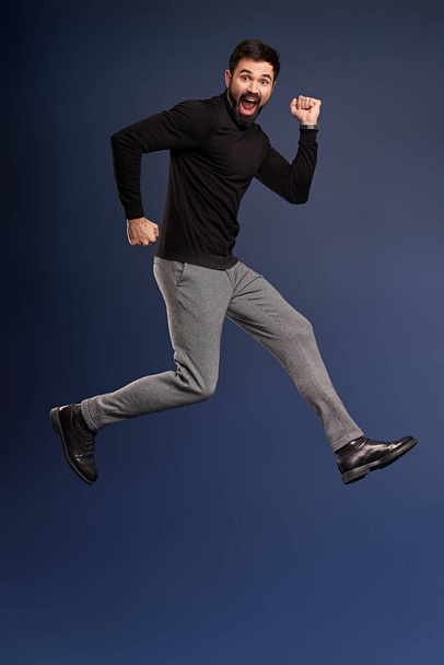 Man in mid-air. Full length of playful young man gesturing and smiling while jumping against Pacific Blue background. - Photo, Image