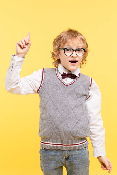 Portrait of emotional schoolboy isolated on yellow background. Happy kid in glasses in vest, bow tie points up with finger to empty spot. Advertising concept of great idea, child's vision. - Photo, Image