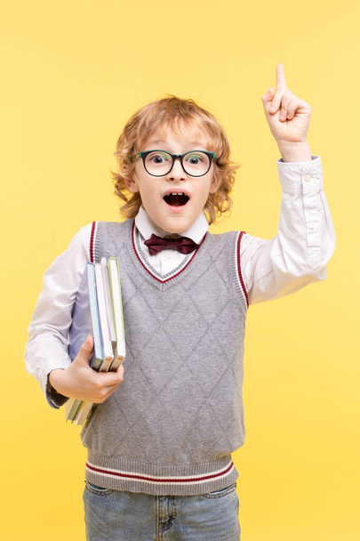 Portrét emocionálního školáka izolovaného na žlutém pozadí. Šťastný chlapec v brýlích ve vestě, s ukazováčkem nahoru, drží knihy. Koncept reklamy na vzdělávací centrum, skvělý nápad, vize. - Fotografie, Obrázek