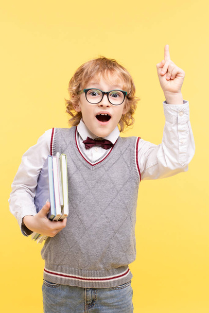 Portrait of an emotional schoolboy isolated on a yellow background. Happy boy in glasses in vest, bow tie points finger up, holds books. Advertising concept for educational center, great idea, vision. - Fotó, kép