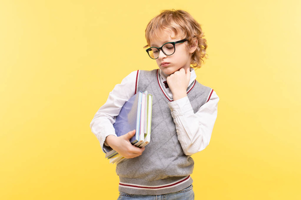 Tired schoolboy with glasses sadly looks at the textbooks at hand. Blond, Caucasian, school uniform. Yellow studio background. Concept of learning problems, bored at school, back to school. Copy space - Foto, immagini