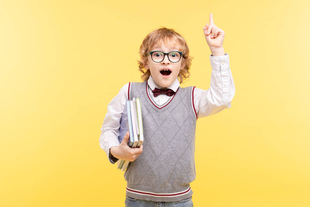Portrait of an emotional schoolboy isolated on a yellow background. Happy boy in glasses in vest, bow tie points finger up, holds books. Advertising concept for educational center, great idea, vision. - Foto, imagen