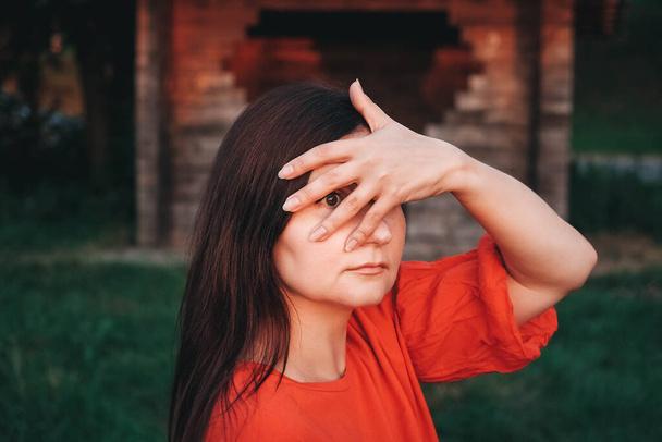 Portrait of a beautiful brunette covering her face with her hand dressed in red clothes stands on the background of a wooden house. Copy, empty space for text. - 写真・画像