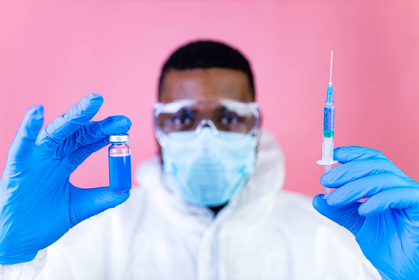 African american man scientist in PPE suite uniform showing medicine liquid vaccine vial bottle coronavirus studio pink wall - Photo, image