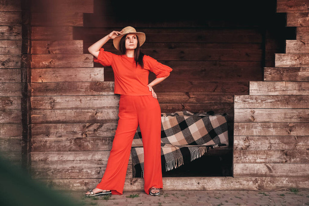 Όμορφη γυναίκα με ψάθινο καπέλο ντυμένη με κόκκινα ρούχα στέκεται στο φόντο ενός ξύλινου σπιτιού. Αντιγραφή, κενό χώρο για κείμενο. - Φωτογραφία, εικόνα