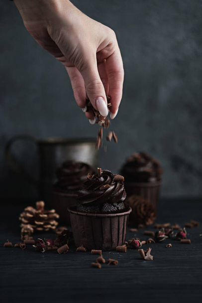 Several muffins or cupcakes with chocolate shaped cream at black table. Festive candle burns on a chocolate cake. A womans hand crumbles grated chocolate onto a cake. - Фото, изображение