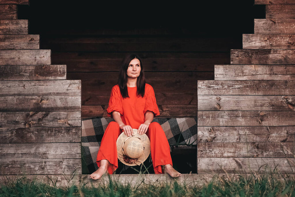 Piękna kobieta w słomkowym kapeluszu ubrana w czerwone ubrania siedząca na tle drewnianego domu. Kopiowanie, puste miejsce na tekst. - Zdjęcie, obraz