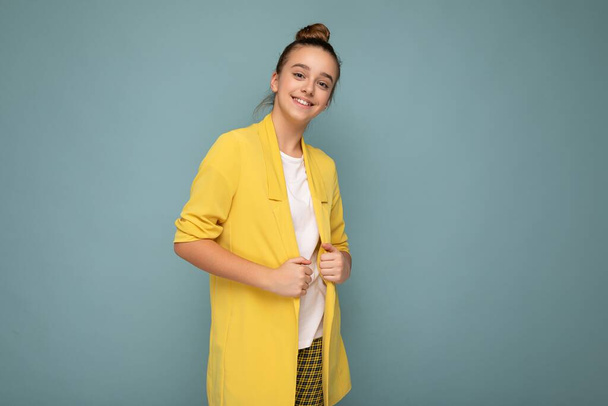 Foto van mooi vrolijk lachend brunette klein meisje in trendy geel jasje en wit t-shirt geïsoleerd over blauwe achtergrond muur kijkend naar camera - Foto, afbeelding