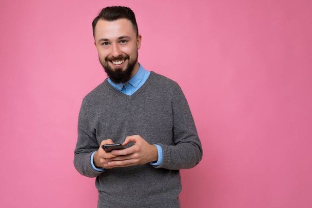Happy smiling handsome good looking brunet bearded young man wearing grey sweater and blue shirt isolated on pink background with empty space holding in hand and using mobile phone communicating - Fotó, kép