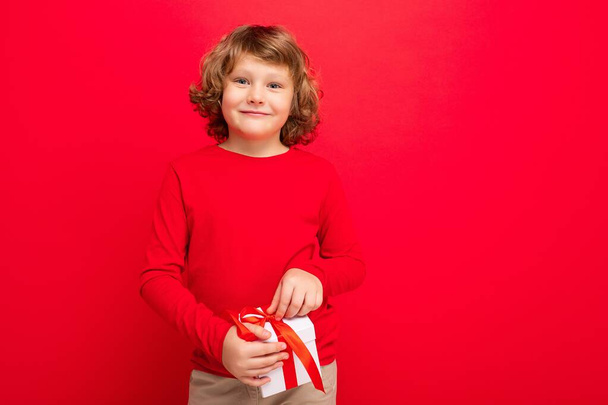 Positivo sorridente ragazzo biondo riccio isolato su parete di sfondo rosso con indosso maglione rosso contenente scatola regalo e guardando la fotocamera - Foto, immagini