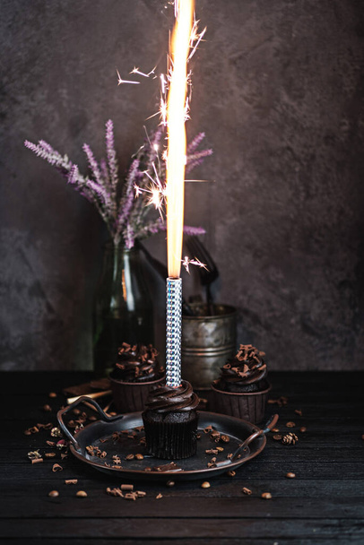 Several muffins or cupcakes with chocolate shaped cream at black table. Festive candle burns on a chocolate cake - Фото, изображение