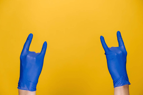 Rock-n-roll y concepto pandémico. Primer plano de las manos masculinas en guantes azules de protección médica ondeando con el gesto de cuernos, aislado en el fondo del estudio amarillo con espacio de copia para la publicidad - Foto, Imagen