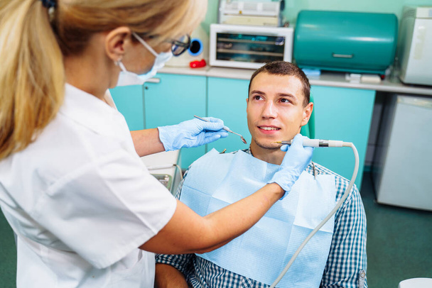 Οδοντιατρική εξέταση δίνεται στον όμορφο άντρα που περιβάλλεται από τον οδοντίατρο και τη βοηθό του. Γυναίκα οδοντίατρος στο οδοντιατρείο μιλάει με τον ασθενή και ετοιμάζεται για θεραπεία. - Φωτογραφία, εικόνα