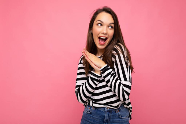 Фото молодой позитивной счастливой удивленной красивой брюнетки женщины с искренними эмоциями в повседневной полосатой пуловер изолирован на розовом фоне с копировальным пространством. Концепция Вау - Фото, изображение