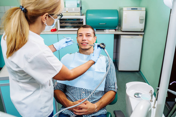 歯科検査は歯科医と助手に囲まれた美しい男に与えられています。歯医者の女性歯科医が患者と話をして治療の準備をしてる. - 写真・画像