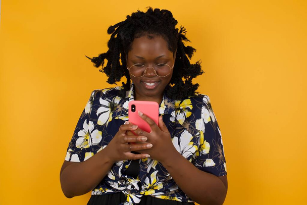 Blonde smiling young african american woman in floral dress demonstrating white teeth using cell phone, messaging, being happy to text with her boyfriend, looking at screen of smartphone. Modern technologies and communication - Photo, Image
