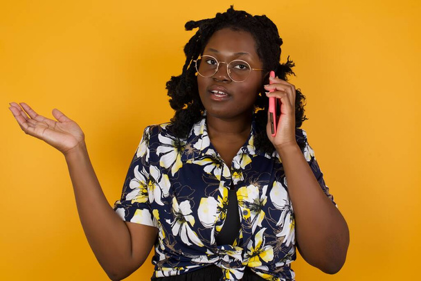 Młoda Afroamerykanka w kwiatowej sukience rozmawiająca przez telefon przez odizolowane tło, zestresowana twarzą w twarz, zszokowana wstydem i niespodziewaną twarzą, zła i sfrustrowana. Strach i zdenerwowanie przez pomyłkę. - Zdjęcie, obraz
