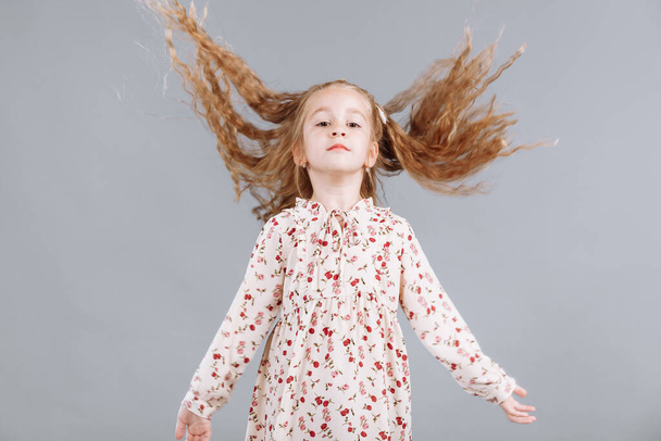 Porträt eines schönen lockigen kleinen Mädchens in einem eleganten floralen Kleid steht, lacht und blickt in die Kamera im Studio auf grauem Hintergrund - Foto, Bild