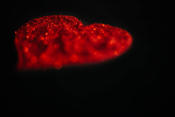 Blurred red heart made of sparkles on a black background. Shimmering dust bokeh symbol - Foto, Imagen