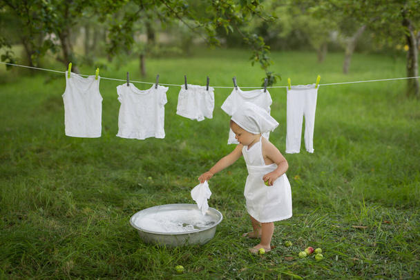 Uma criança lava alegremente suas roupas na bacia. Pulverize água e espuma da roupa lavada. Menina de vestido branco pendurado lavandaria molhada para secar. Lavagem em uma bacia vintage. - Foto, Imagem