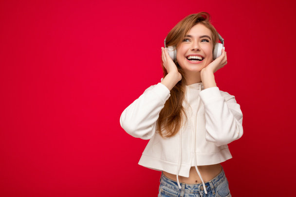 Photo of beautiful happy smiling young blonde woman wearing white hoodie isolated over colourful background wall wearing white wireless bluetooth earphones listening to cool music and enjoying looking - Zdjęcie, obraz