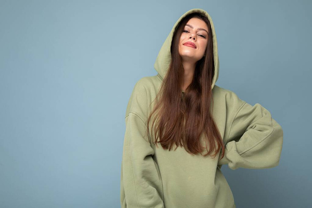 Young beautiful woman looking at camera. Trendy girl in casual hipster hoodie clothes. Positive female shows facial emotions. Funny model isolated on blue background with free space for text - Foto, Imagen