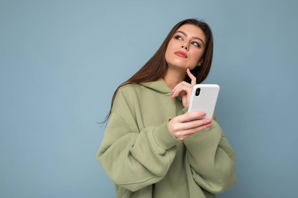 Pretty adult brunette woman wearing stylish green hoodie using mobile phone writing sms isolated on background looking up and thinking - Foto, afbeelding