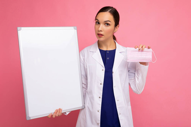 Vrouwelijke persoon arts in een witte medische jas houden blanco boord met kopieerruimte voor tekst en beschermende masker geïsoleerd op roze achtergrond. Epidemisch concept - Foto, afbeelding