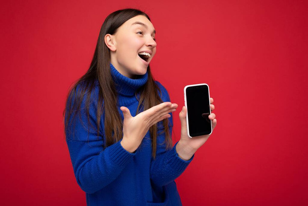 Foto van mooie jonge brunette vrouw met casual blauwe trui geïsoleerd over rode achtergrond met lege ruimte in de hand mobiele telefoon en toont smartphone met leeg scherm voor mockup - Foto, afbeelding