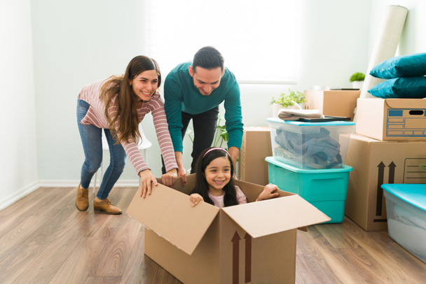 Glimlachende jonge vrouw en man duwen een klein meisje in een kartonnen doos. Speelse latijnse familie die plezier hebben in de woonkamer tijdens het inpakken van hun meubels en dingen  - Foto, afbeelding