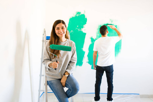 Handig vrouwtje dat de muren schildert met een verfroller langs haar liefhebbende man. Getrouwd echtpaar renovatie en herinrichting van hun nieuwe huis - Foto, afbeelding