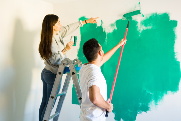 Latynoska dziewczyna po dwudziestce wskazująca na swojego poręcznego chłopaka. Miejsce, które przegapił, by namalować na świeżo pomalowanej ścianie.   - Zdjęcie, obraz
