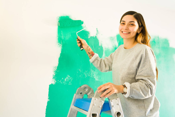 Портрет веселой женщины 20 лет, улыбающейся, держа в руках ролик с краской. Независимая женщина красит стены своей новой квартиры или дома - Фото, изображение