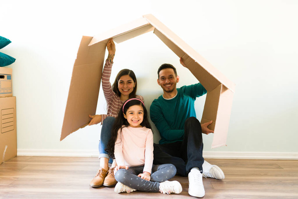 Spaanse familie houdt een kartonnen doos boven hun hoofd om het dak van een huis te vormen. Mooie moeder, vader en dochter zitten op de vloer op verhuisdag - Foto, afbeelding