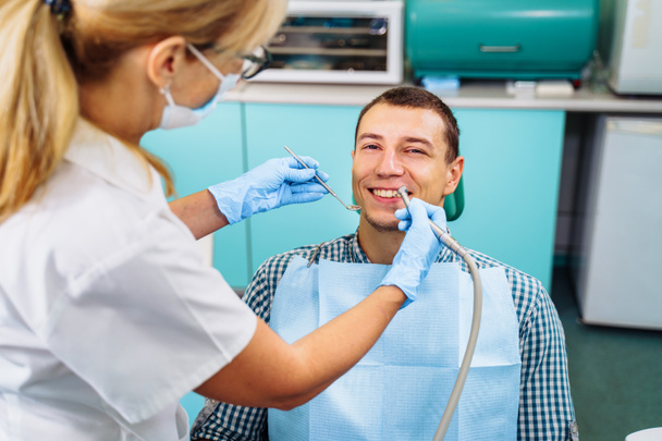 A young, attractive European-looking guy came to the dentist. Sits in a dental chair. The dentist bent over him and prepares to treat problem teeth. Happy patient and dentist concept. - Photo, Image