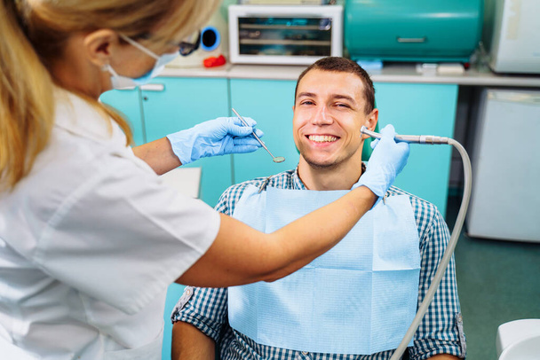 Ένας νεαρός, ελκυστικός Ευρωπαίος ήρθε στον οδοντίατρο. Κάθεται σε μια οδοντιατρική καρέκλα. Ο οδοντίατρος έσκυψε πάνω του και ετοιμάζεται να θεραπεύσει τα προβληματικά δόντια. Ευτυχής ασθενής και οδοντίατρος έννοια. - Φωτογραφία, εικόνα