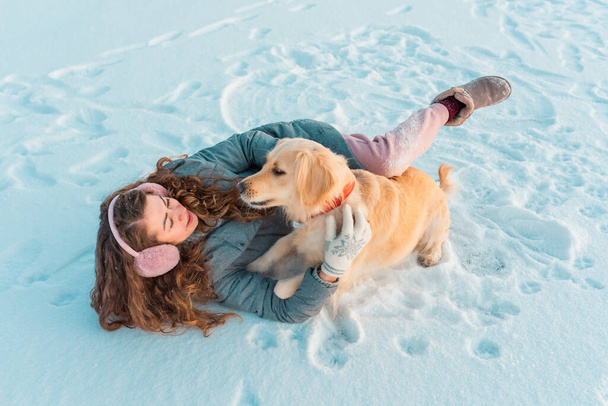 Μακρύ σγουρό νεαρό κορίτσι σε ροζ ωτοασπίδες χειμώνα και λευκά γάντια παίζει με το σκύλο της στο κρύο παγωμένο ποτάμι χειμώνα καλυμμένο με χιόνι. Χιονίζει χειμώνα έννοια. - Φωτογραφία, εικόνα