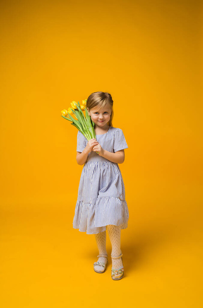 una niña con un vestido a rayas se levanta con un ramo de tulipanes amarillos sobre un fondo amarillo con espacio para el texto - Foto, Imagen