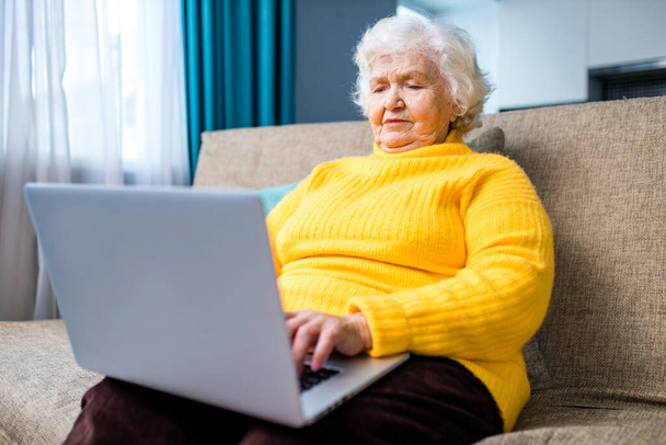 aged mature grey haired woman in yellow sweater sitting on sofa with laptop in living room - Foto, Imagen