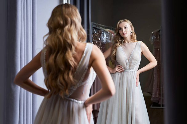 Young beautiful blonde girl wearing a full-length silver white chiffon prom ball gown decorated with sparkles and sequins. Model in front of mirror in a fitting room at dress hire service. - Фото, изображение