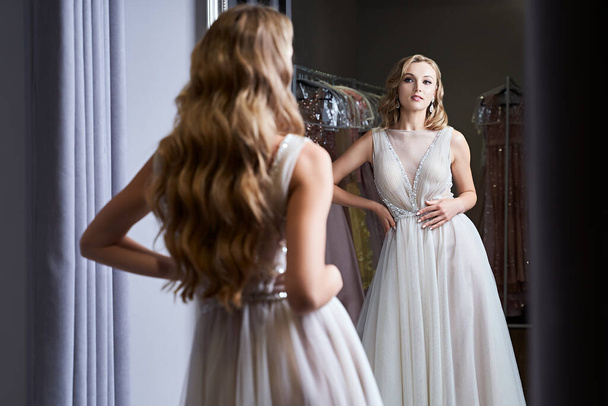 Young beautiful blonde girl wearing a full-length silver white chiffon prom ball gown decorated with sparkles and sequins. Model in front of mirror in a fitting room at dress hire service. - Фото, изображение
