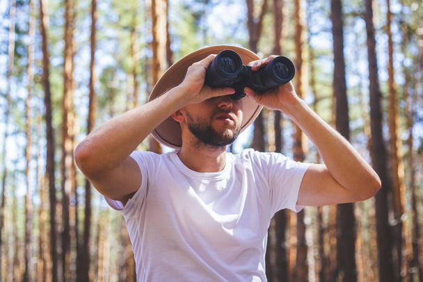 Man in a hat looks through binoculars during a camping trip. Hike in the mountains, forest. - Foto, imagen