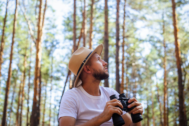 Man in a hat holds binoculars during a camping trip. Hike in the mountains, forest. - Photo, Image