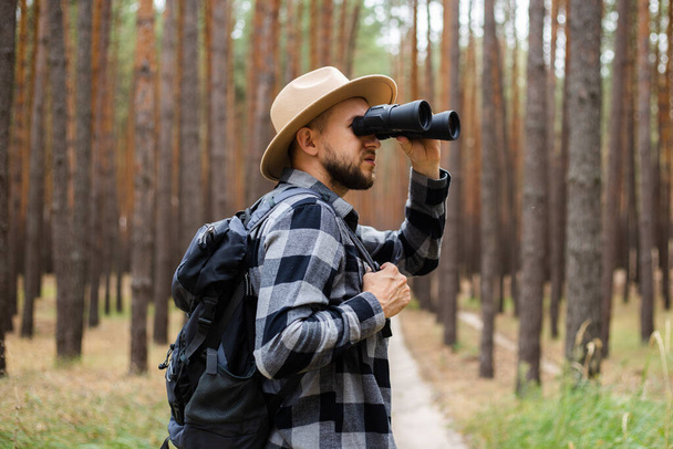 Man met een hoed en een geruit shirt houdt een verrekijker vast en loopt door het bos. - Foto, afbeelding