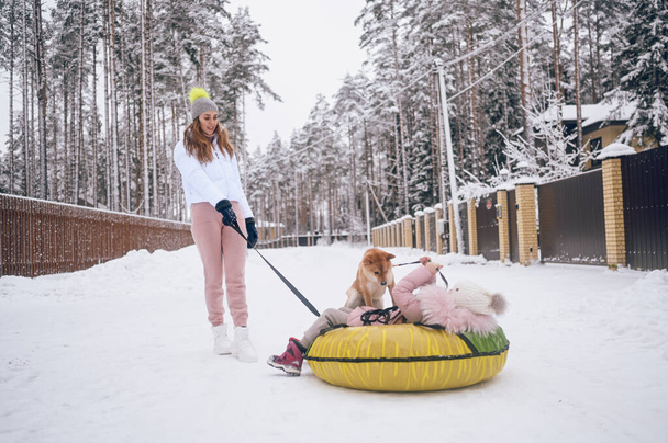 Szczęśliwa matka i mała słodka dziewczyna w różowej ciepłej bieliźnie spacerując o zabawy przejażdżki nadmuchiwane rury śnieżnej z czerwonym shiba inu pies w śniegu białej zimie wsi na zewnątrz. Rodzinne zajęcia sportowe - Zdjęcie, obraz