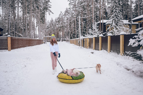 Щаслива мати і маленька мила дівчинка в рожевому теплому одязі гуляють надувні снігові пробки з червоною собакою шиба в сніжно-білій зимовій сільській місцевості на відкритому повітрі. Сімейна спортивна відпустка
 - Фото, зображення