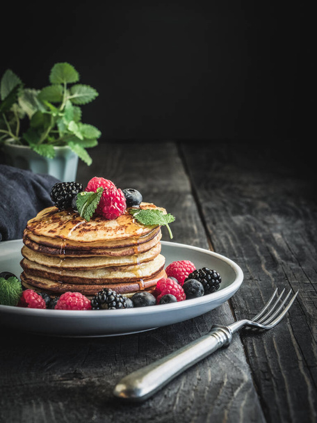 Pancakes with berries - blueberries, raspberries and blackberries and mint leaves on plate and vintage wooden table. Jar of honey on background. - Photo, Image