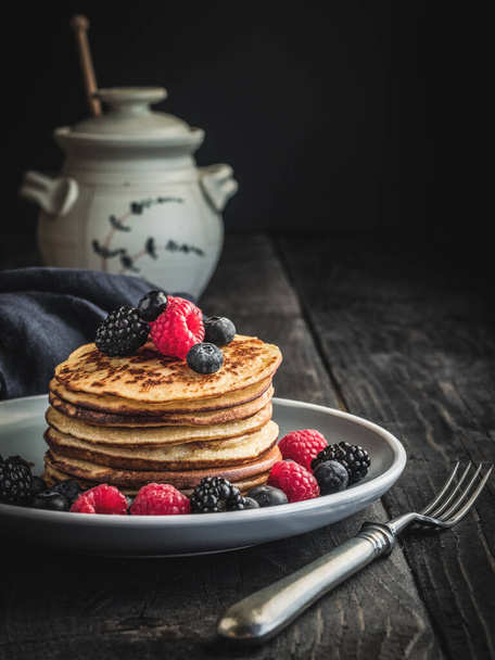 Pancakes with fresh berries - blueberries, raspberries and blackberries on plate and vintage wooden table. Jar of honey on blurry background. - Photo, Image