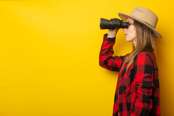 Giovane donna in cappello e camicia a quadri guardando attraverso il binocolo su sfondo giallo. - Foto, immagini
